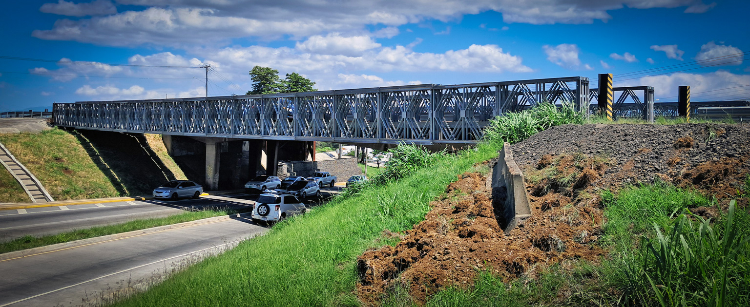 Puente vehicular metálico tipo Bailey sobre carretera, con estructura de acero gris y paso de vehículos por debajo, rodeado de vegetación y cielo despejado.