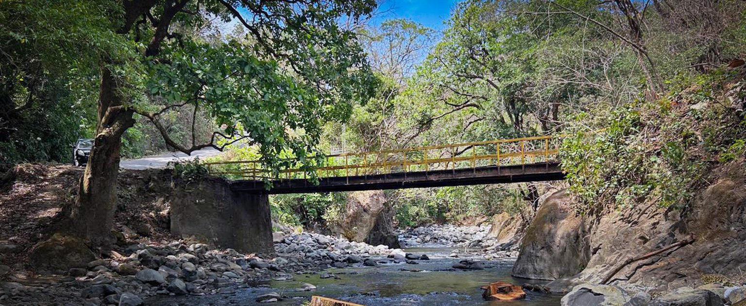 Puente vehicular angosto con barandas amarillas que cruza el río Barranquilla, rodeado de vegetación densa y rocas, con un cauce de aguas poco profundas y una carretera visible en uno de los extremos.