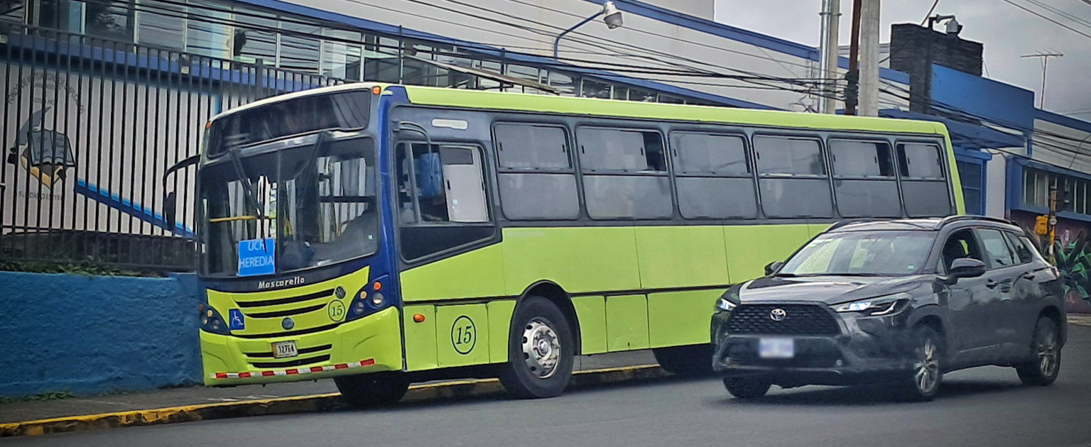 Autobús de transporte público de color verde y azul circulando por una vía urbana, junto a un vehículo particular. Al fondo se observan instalaciones comerciales, aceras y cableado aéreo