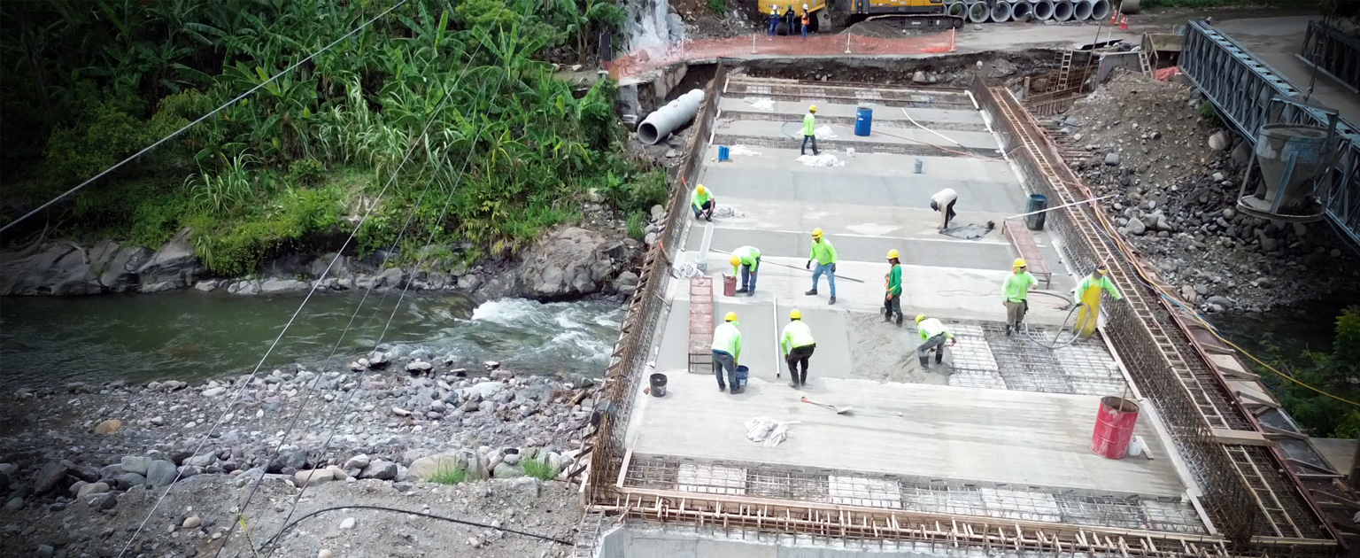 Puente sobre el río Guacimal en construcción, con trabajadores colocando concreto en la losa mientras el cauce del río y vegetación se observan a un costado.