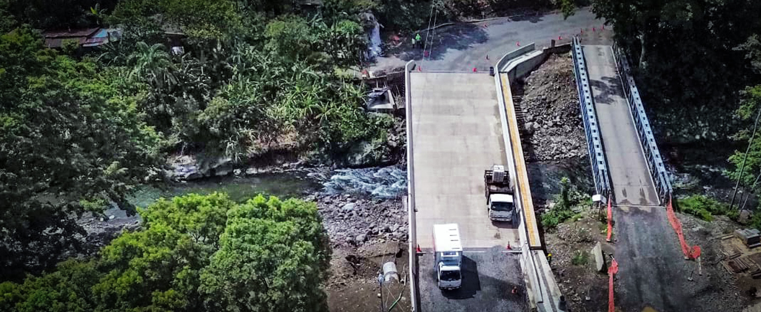 Nuevo puente sobre el río Guacimal junto al antiguo puente metálico, rodeado de vegetación y con tránsito vehicular sobre la nueva estructura.