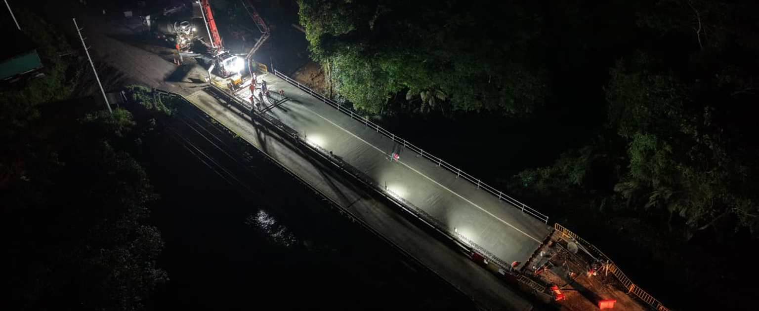Vista aérea nocturna de los trabajos de construcción del puente sobre el río Las Haciendas. La losa del nuevo puente se observa recién colocada e iluminada por reflectores, mientras maquinaria especializada y personal de obra realizan labores de fundido de concreto. El puente está rodeado de vegetación densa y señalización temporal de construcción.