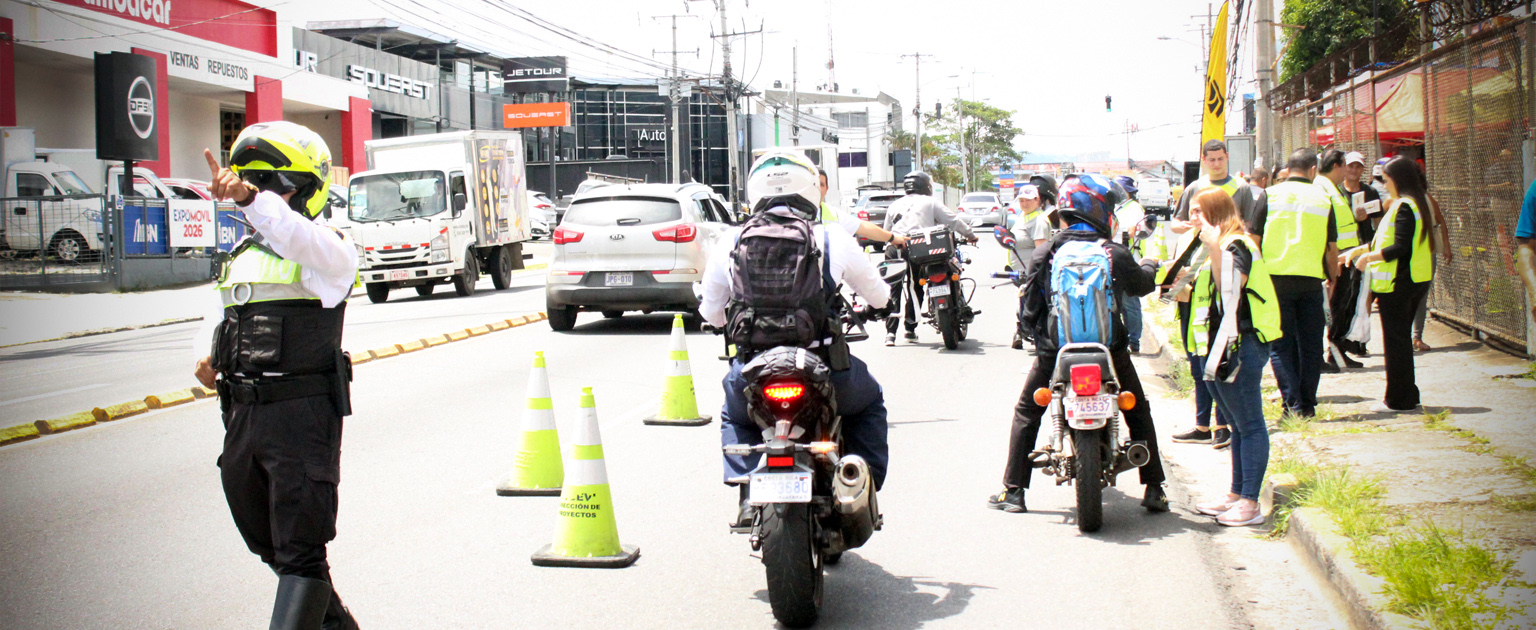 Operativo de control vial en carretera urbana, con oficiales regulando el tránsito de motocicletas mediante conos, mientras varias personas observan desde la acera y circulan vehículos al fondo.