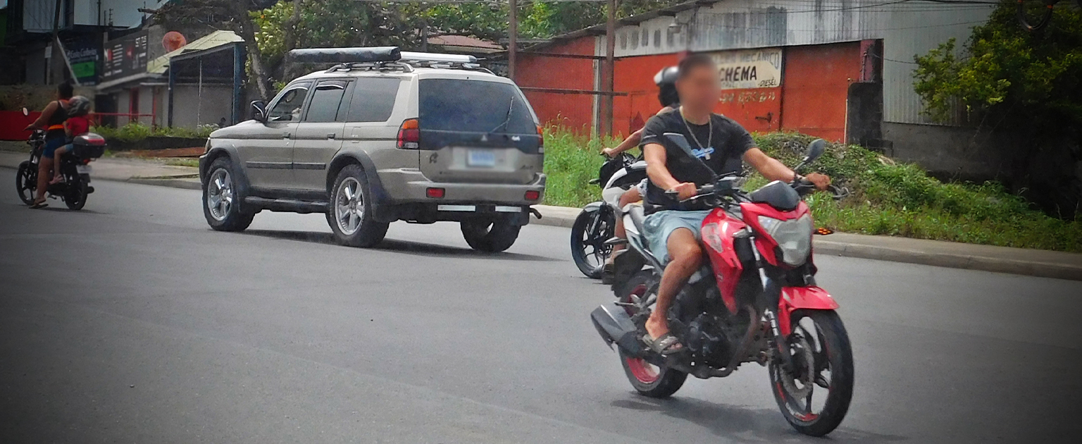 Fotografía que muestra dos motociclistas sin casco en una carretera a dos carriles donde circulan otros vehículos