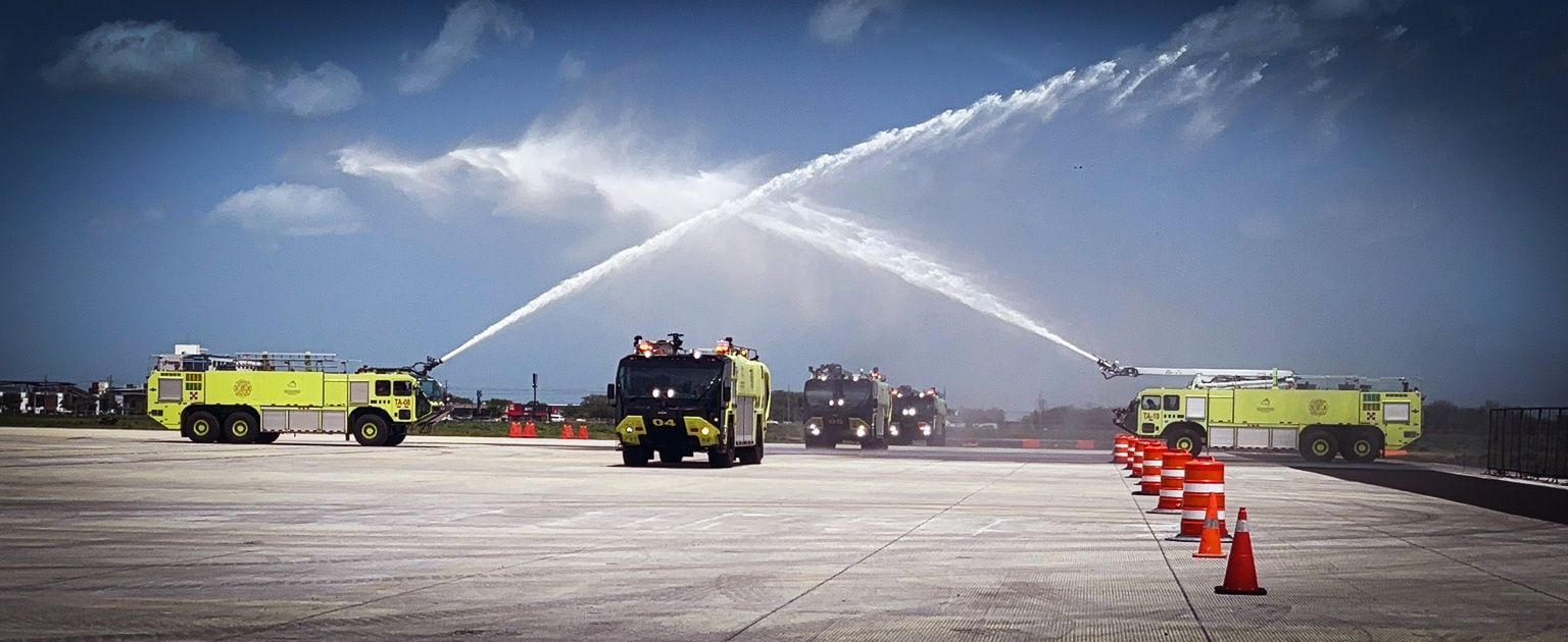 Pista del Aeropuerto Internacional Daniel Oduber, con camiones de bomberos aeroportuarios realizando arco de agua sobre la superficie, mientras se observan conos de señalización y cielo parcialmente nublado al fondo.