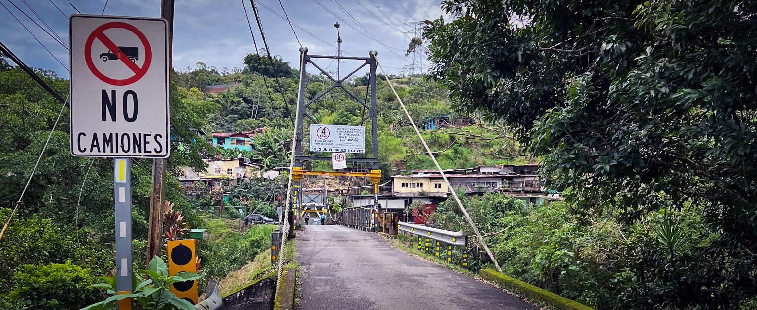 Actual puente sobre el río Palomo con señal de prohibido camiones, rodeado de vegetación y viviendas, con circulación controlada