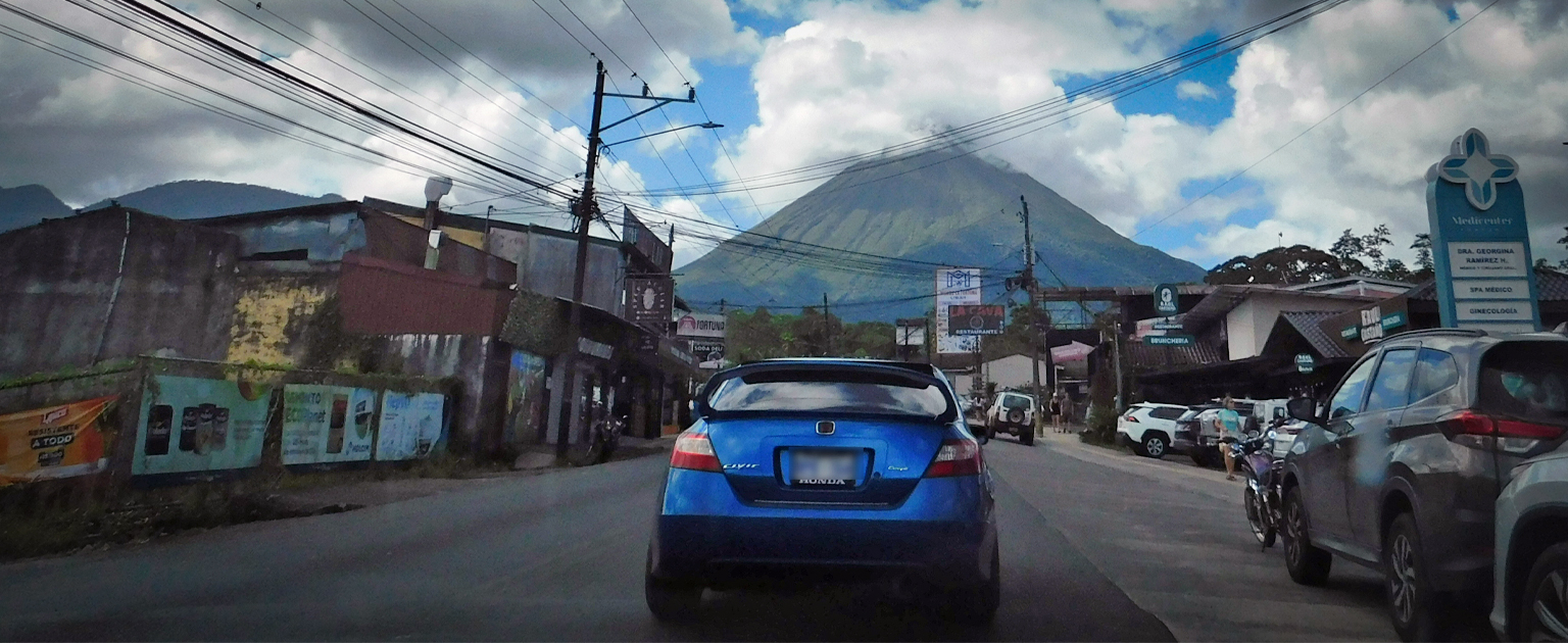 Vista de una calle urbana en La Fortuna, con vehículos circulando y comercios a ambos lados de la vía. Al fondo se aprecia el volcán Arenal elevándose sobre el paisaje, parcialmente rodeado de nubes, mientras postes y tendido eléctrico enmarcan la escena