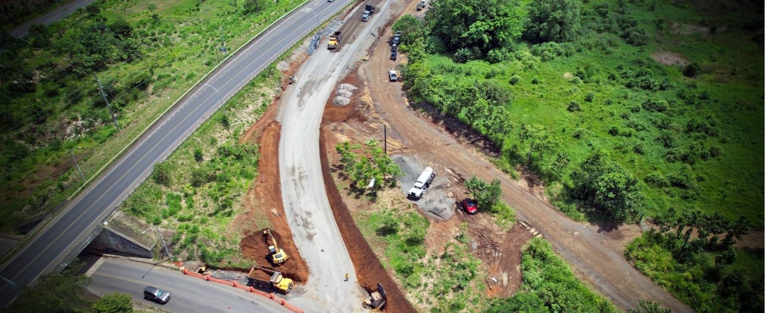 En la fotografía se observa una vista aérea de una obra vial en construcción. A la izquierda, se extiende una carretera pavimentada, mientras que en el centro y a la derecha de la imagen se despliega una nueva vía en proceso de pavimentación. La zona en construcción está rodeada de áreas verdes y árboles. Varias máquinas de construcción, como excavadoras y camiones, se encuentran trabajando sobre el terreno, nivelando y preparando el espacio para la ampliación de la carretera.