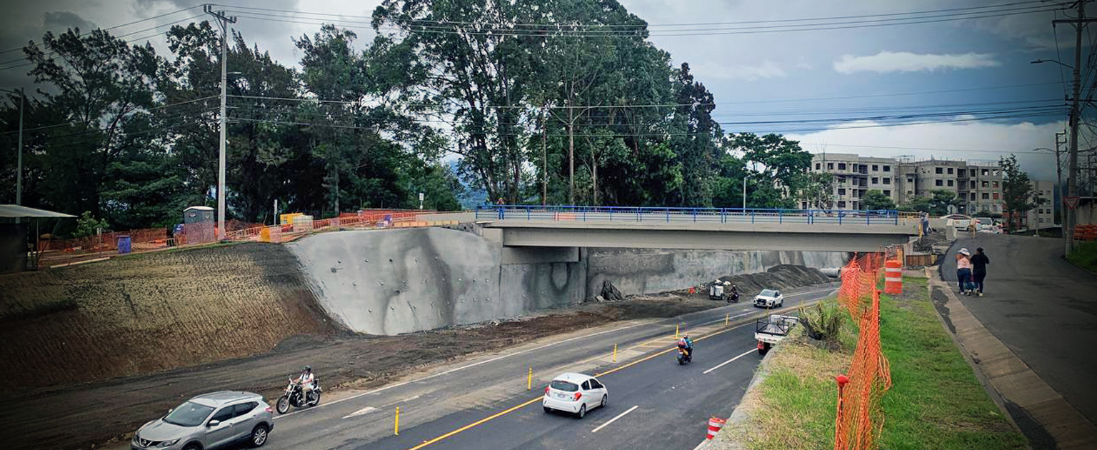 La estructura elevada está flanqueada por barreras de contención y señalización naranja que delimita el área en obra. Abajo, se observa la calle con varios vehículos en circulación y un motociclista. Al fondo, se ven edificios residenciales y una zona de árboles frondosos bajo un cielo parcialmente nublado. A la derecha, dos peatones caminan por la acera cercana al área de construcción."