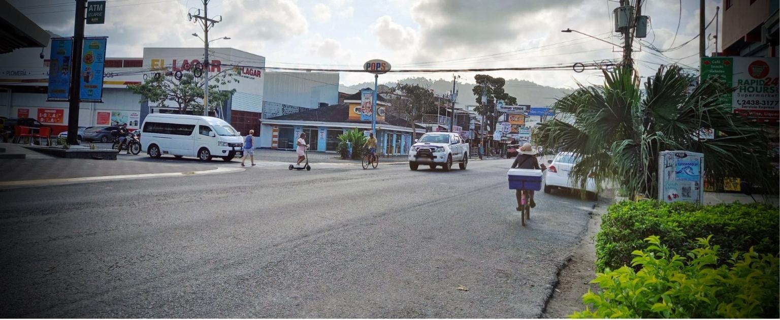 Cruce la pops en Jacó. Una persona en bicicleta, otra en es scouter y muchos vehículos en carretera. 