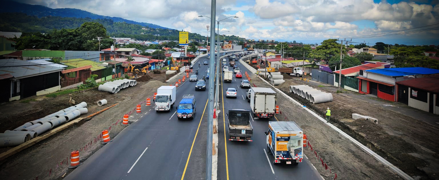 En el centro, una carretera principal de cuatro carriles con alto flujo vehicular es flanqueada por áreas de construcción delimitadas por conos anaranjados. A ambos lados de la vía se observan pilas de tubos de concreto, indicativos de trabajos de alcantarillado o drenaje pluvial.