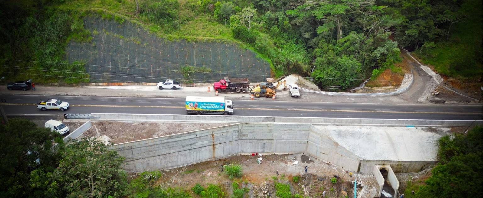 La fotografía muestra una obra vial en una zona montañosa rodeada de vegetación densa. En el centro, una carretera asfaltada cuenta con vehículos estacionados y maquinaria pesada, como una retroexcavadora y un camión, que trabajan en la construcción o mantenimiento de la vía