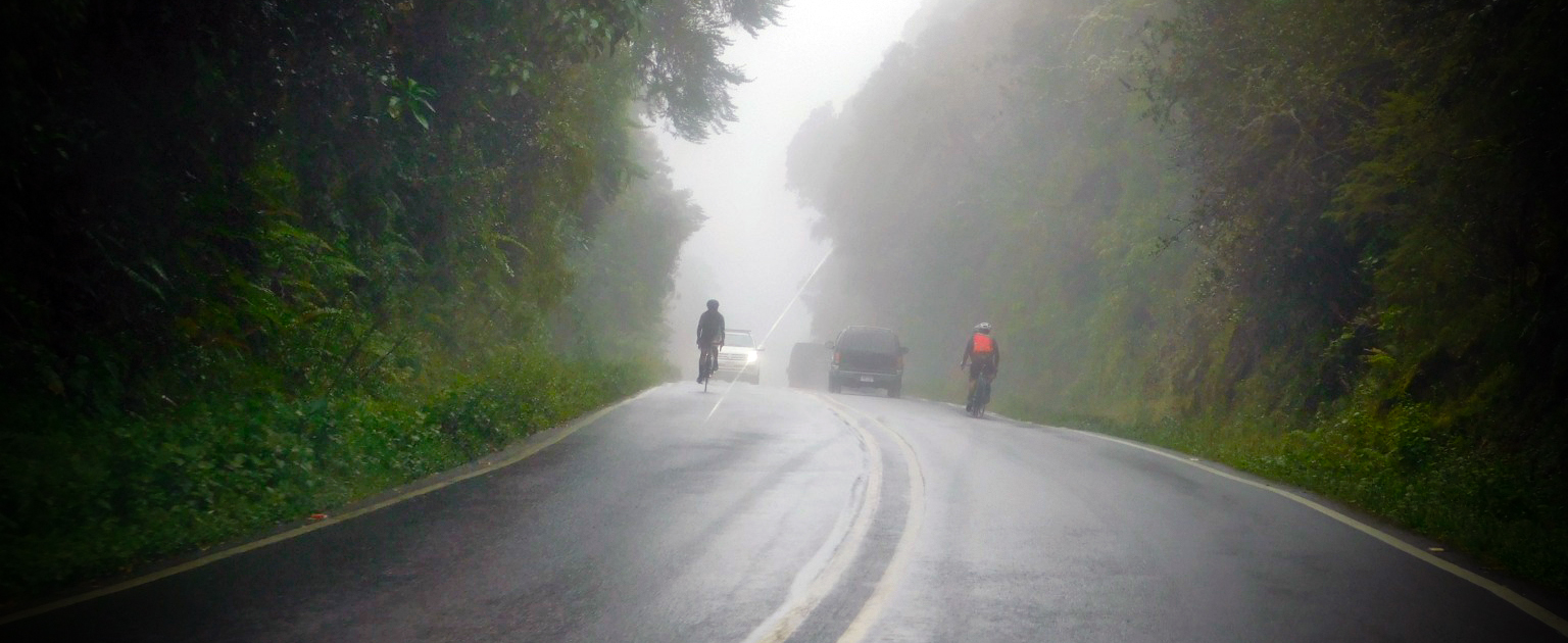 Ciclistas en carretera que se encuentra en medio de dos montañas. Se ve nublado.