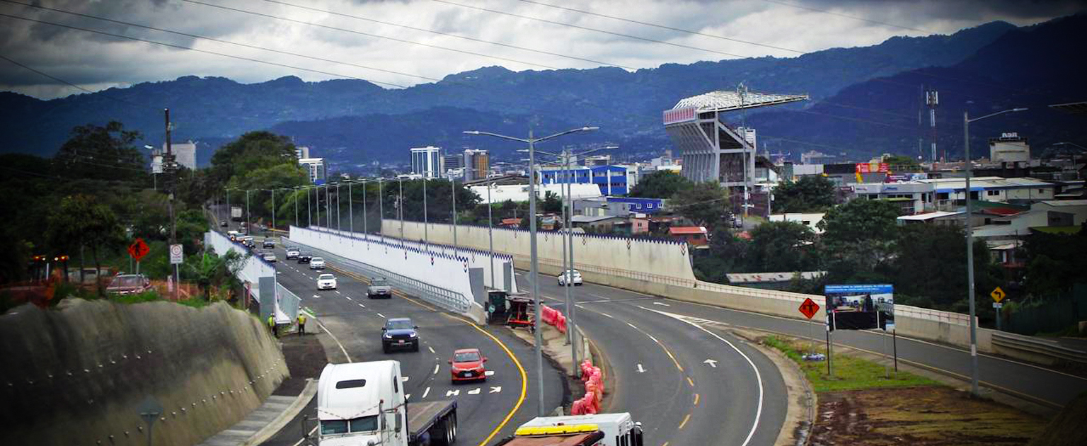 Fotografía aérea del nuevo puente y vehículos transitando. A la derecha se observa el Estadio Ricardo Saprissa y edificios de la ciudad en el fondo, enmarcados por montañas bajo un cielo nublado. 