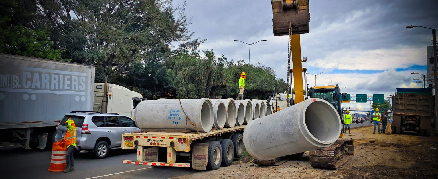 Un camión de plataforma transporta tubos de concretos, mientras que una máquina retroexcavadora realiza la descarga con ayuda de cadenas y personal técnico. Varios trabajadores con equipo de seguridad, supervisan y colaboran en la operación. 