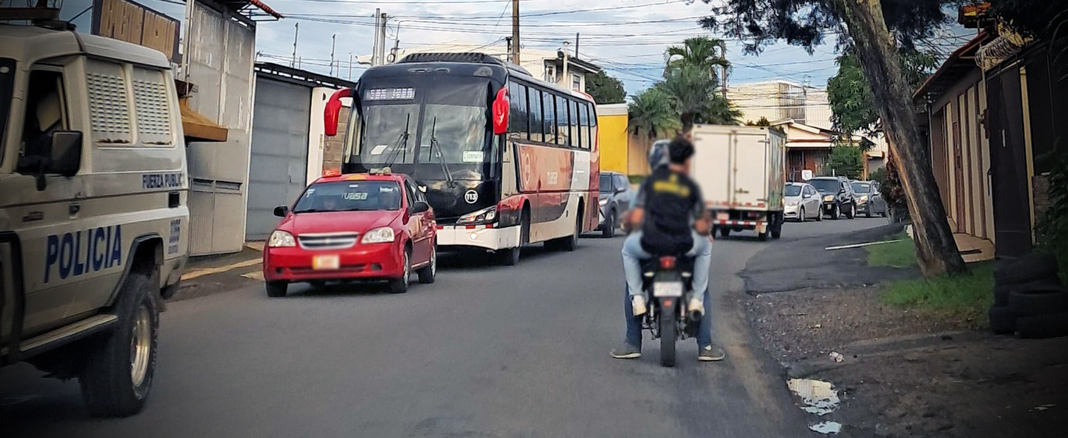 Fotografía que muestra una carretera con varios vehículos circulando y a la derecha dos personas en moto, una de ellas sin casco