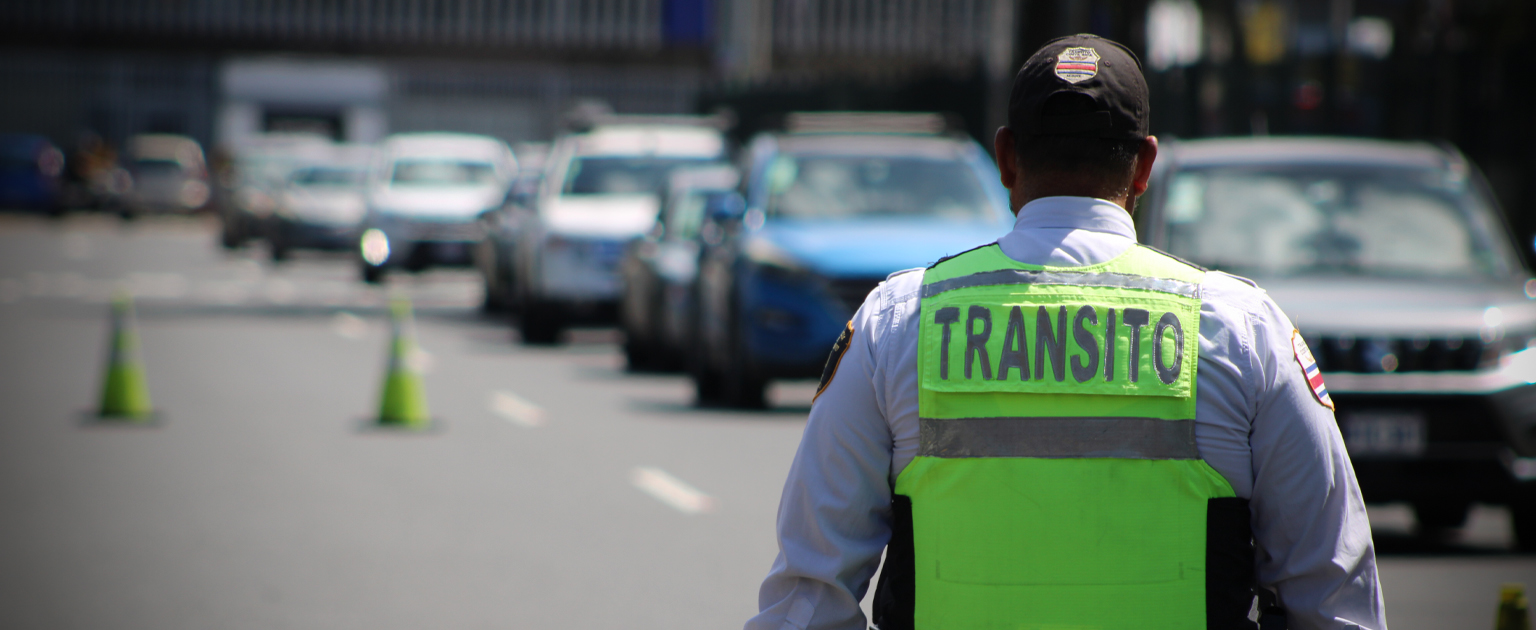 Oficial de tránsito con chaleco reflectante verde supervisa la circulación de vehículos en una carretera de varios carriles. Al fondo se observa una fila de automóviles y conos de señalización que delimitan el flujo vehicular