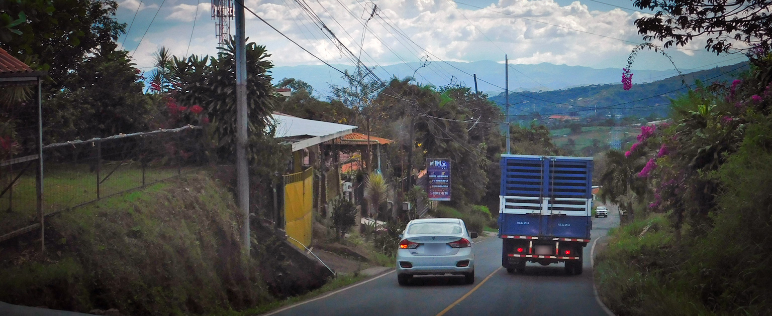 Carretera de dos carriles en una zona rural y montañosa, con un vehículo liviano realizando una maniobra de adelantamiento indebido a un camión. A ambos lados de la vía se observan viviendas, vegetación abundante, postes y tendido eléctrico. Al fondo se distinguen colinas y montañas bajo un cielo parcialmente nublado.