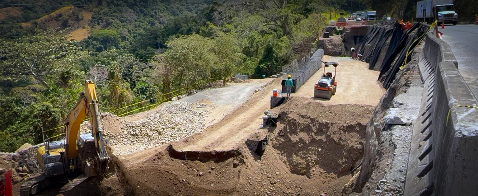 Vista de las intervenciones viales en el sector de Cambronero. En primer plano se observan trabajos de excavación y conformación de terreno con maquinaria pesada, mientras operarios realizan labores de estabilización y mejora de la infraestructura. A la derecha se aprecia la carretera en servicio, protegida por barreras de concreto, y al fondo estructuras de contención para reforzar el talud. El proyecto se desarrolla en una zona montañosa rodeada de abundante vegetación.