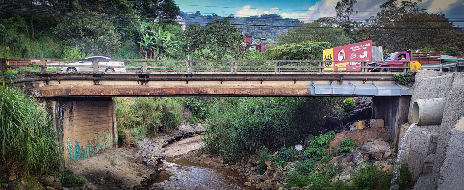 Puente sobre el río Cañas, con estructura vehicular de concreto y paso de vehículos, ubicado en un entorno urbano con vegetación, cables eléctricos y un sistema de drenaje lateral visible