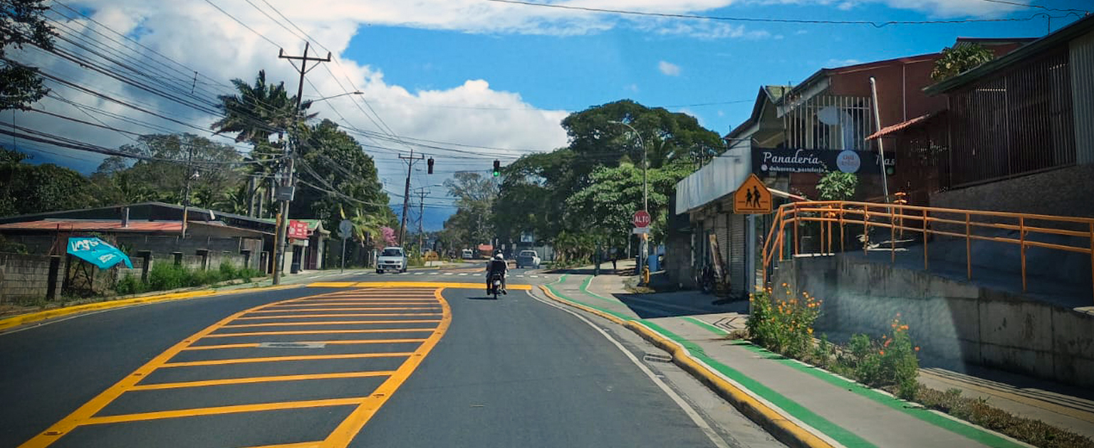 Proyecto conocido como calle completa en Alajuela, se muestra una calle de doble vía en zona urbana con señalización vial recién pintada en color amarillo, acera con ciclovía verde a la derecha, una motocicleta circulando al centro y semáforo en verde al fondo; a los lados hay viviendas, comercios y vegetación.