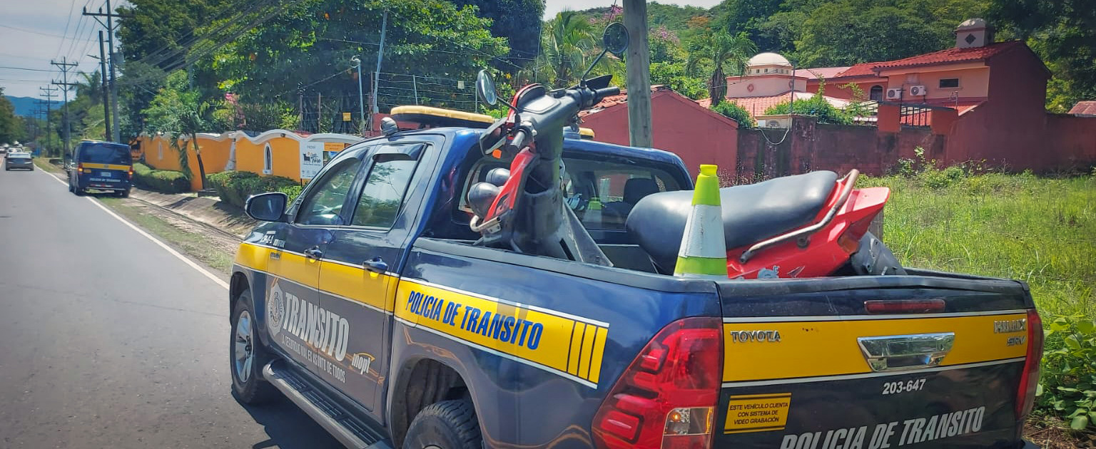 Patrulla tipo pick up de la Policía de Tránsito estacionado a la orilla de una carretera, con una motocicleta decomisada y un cono de seguridad en el cajón, en un entorno urbano con viviendas y vegetación cercana