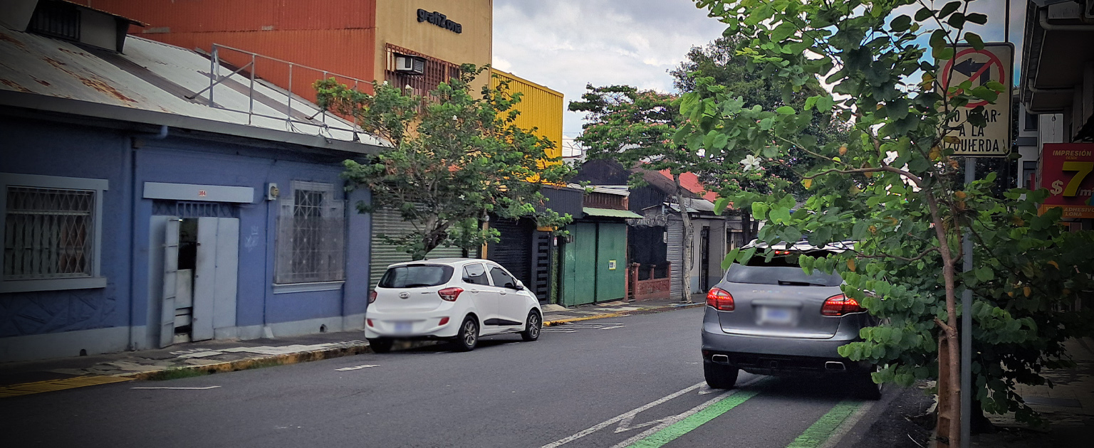 Vehículo estacionado sobre la ciclovía en una calle urbana, junto a una señal de No virar a la izquierda, muestra otros automóviles y viviendas alrededor.