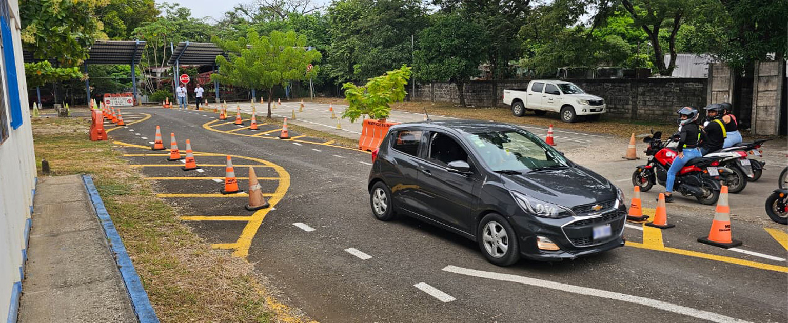 Vehículo liviano circula por el circuito interno destinado a la  prueba práctica en Nicoya. A un costado de la vía se observan dos motocicletas con sus ocupantes y, al fondo, una zona de estacionamiento rodeada de árboles y vegetación. La pista cuenta con demarcación horizontal para guiar las maniobras de los conductores.