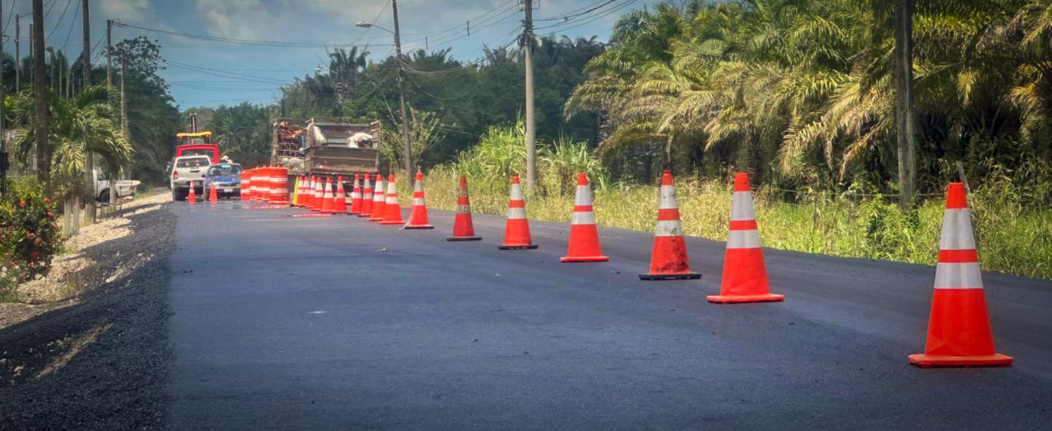 Conos de señalización delimitan un tramo de carretera recién asfaltada mientras se desarrollan trabajos viales. Al fondo se observan vehículos y maquinaria de apoyo.