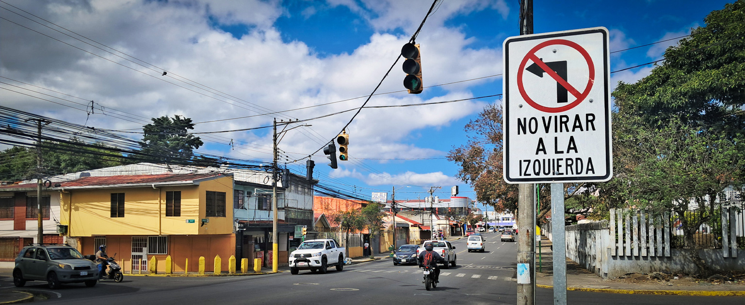 Fotografía que muestra un cruce semaforizado y en primer plano una señal de tránsito que indica no virar a la izquierda