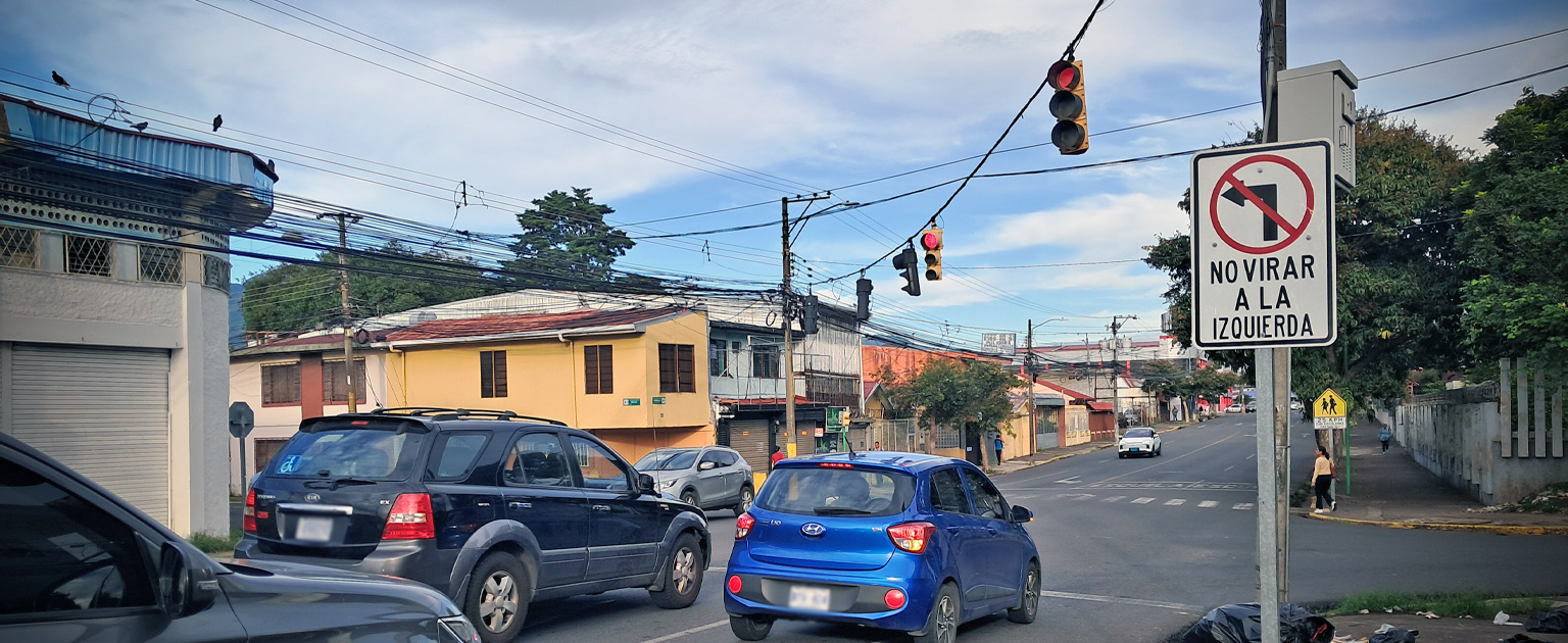 Vista urbana de una intersección regulada por semáforos, con varios vehículos detenidos ante la luz roja. En primer plano destaca una señal vertical de tránsito con la indicación “No virar a la izquierda”. La vía está rodeada de edificaciones residenciales y comerciales, aceras peatonales, postes de servicios públicos y abundante cableado aéreo. La imagen ilustra una medida de regulación vial para ordenar los movimientos de giro y mejorar la seguridad en el cruce.