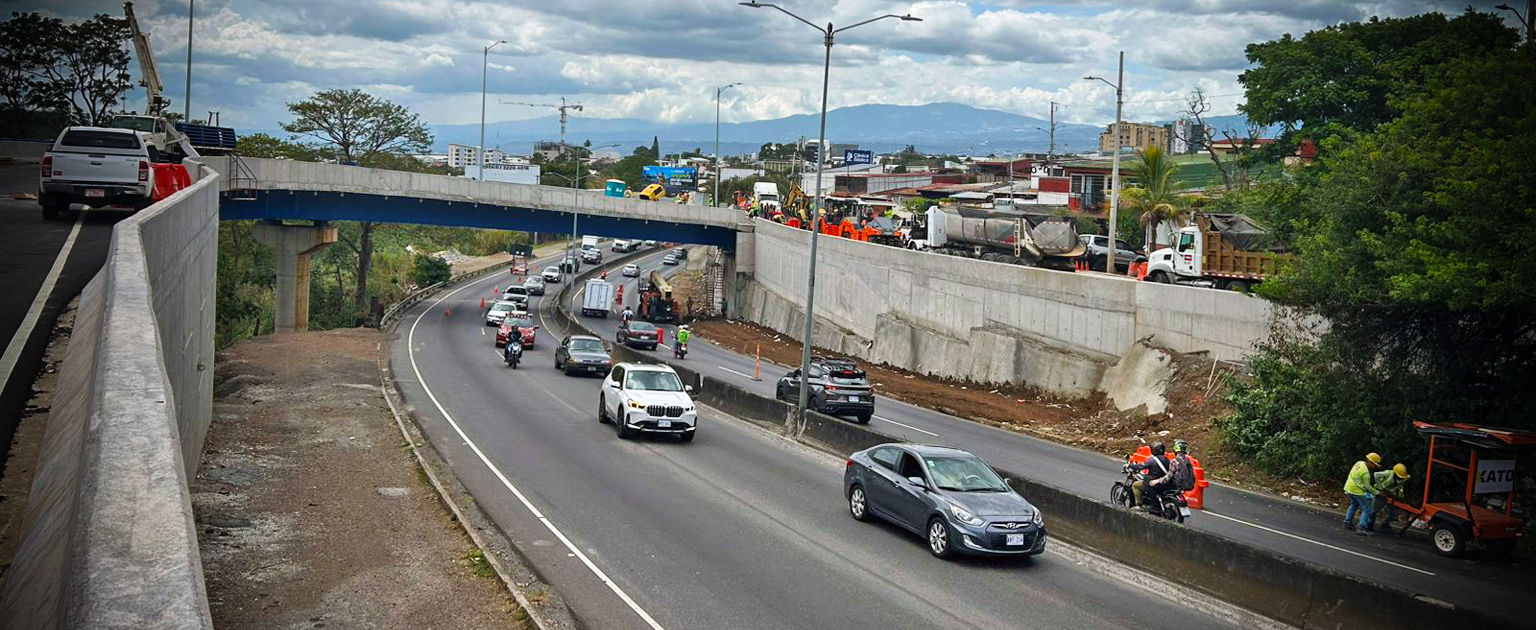 Paso a desnivel de Hatillos 7 y 8 en carretera con tránsito en ambos sentidos, donde circulan vehículos livianos y motocicletas; en el nivel superior y lateral derecho se observan trabajos viales con maquinaria y personal de obra, mientras al fondo se distinguen zonas urbanas y montañas bajo cielo parcialmente nublado.