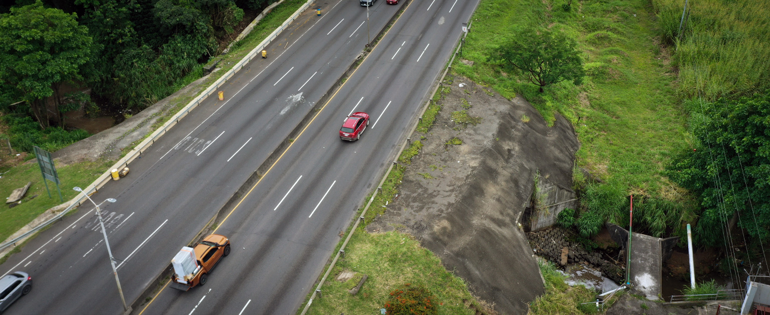 Vista aérea de la Ruta Nacional uno a cuatro carriles con circulación vehicular moderada. A un lado de la vía se observa el cauce de quebrada La Guaria y la alcantarilla.