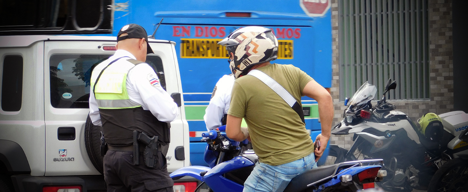 Control vial en zona urbana: oficial de tránsito detiene a motociclista con casco junto a varios vehículos, incluyendo una patrulla y una motocicleta policial; al fondo se observa un autobús