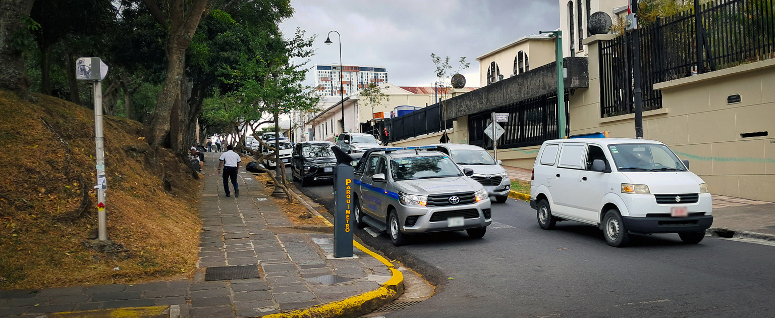 Calle urbana en las cercanías de la Asamblea Legislativa con tráfico vehicular, acera con peatones y parquímetro en una zona arbolada