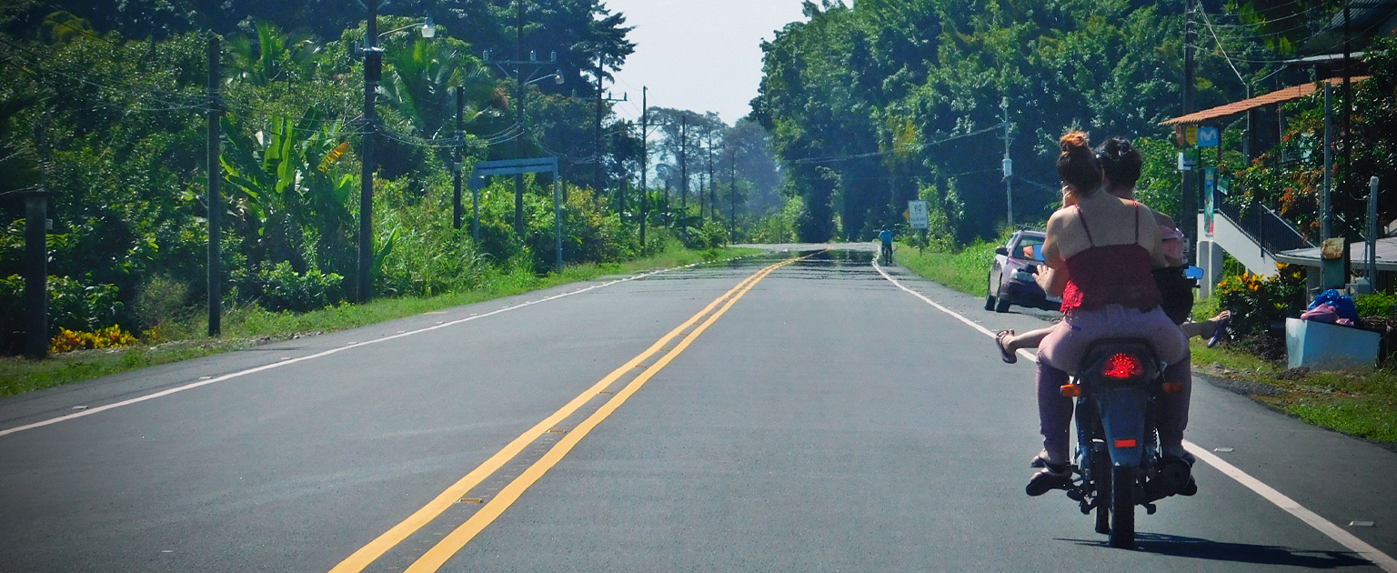  Motocicleta con tres personas a bordo transita por una carretera asfaltada en una zona rural sin casco ni prendas reflectivas. La imagen ilustra una situación de riesgo