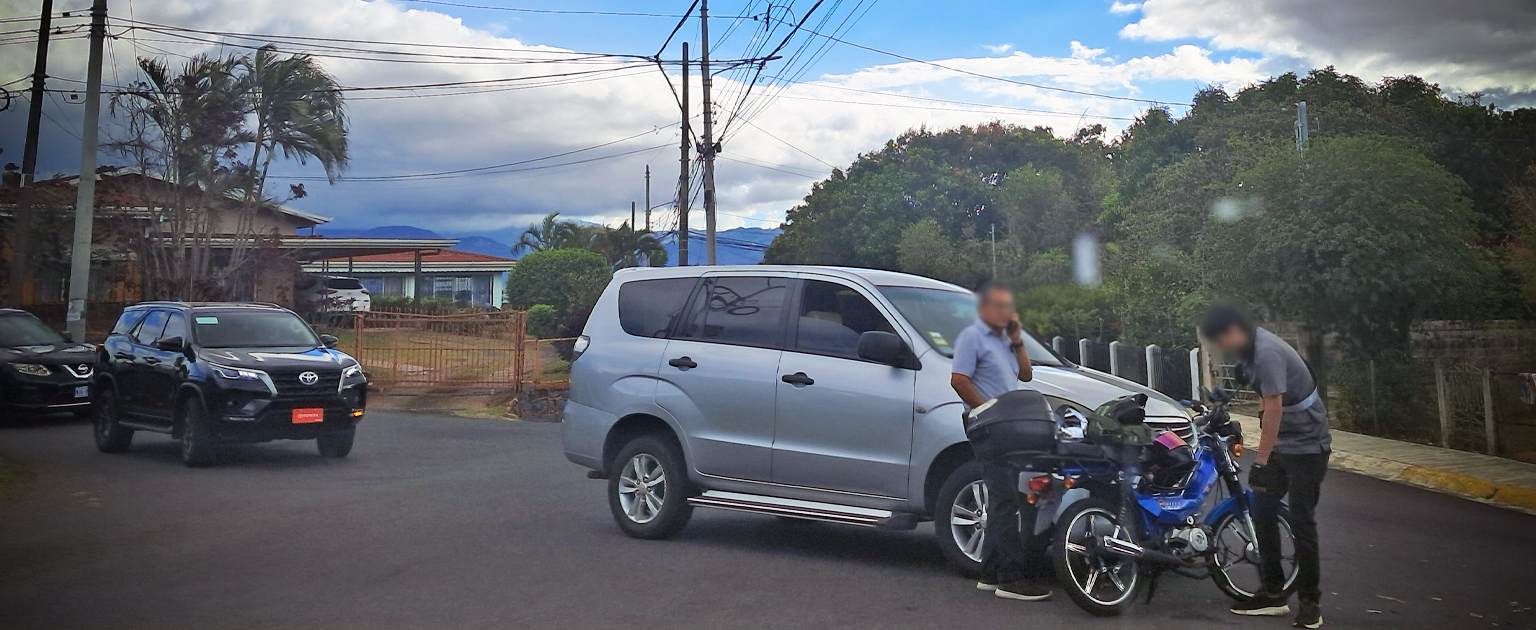 Escena de accidente de tránsito en vía urbana: una motocicleta detenida junto a un vehículo tipo SUV gris, con dos personas de pie cerca revisando o conversando, mientras otros vehículos esperan en la carretera; al fondo se observan viviendas, postes eléctricos y árboles