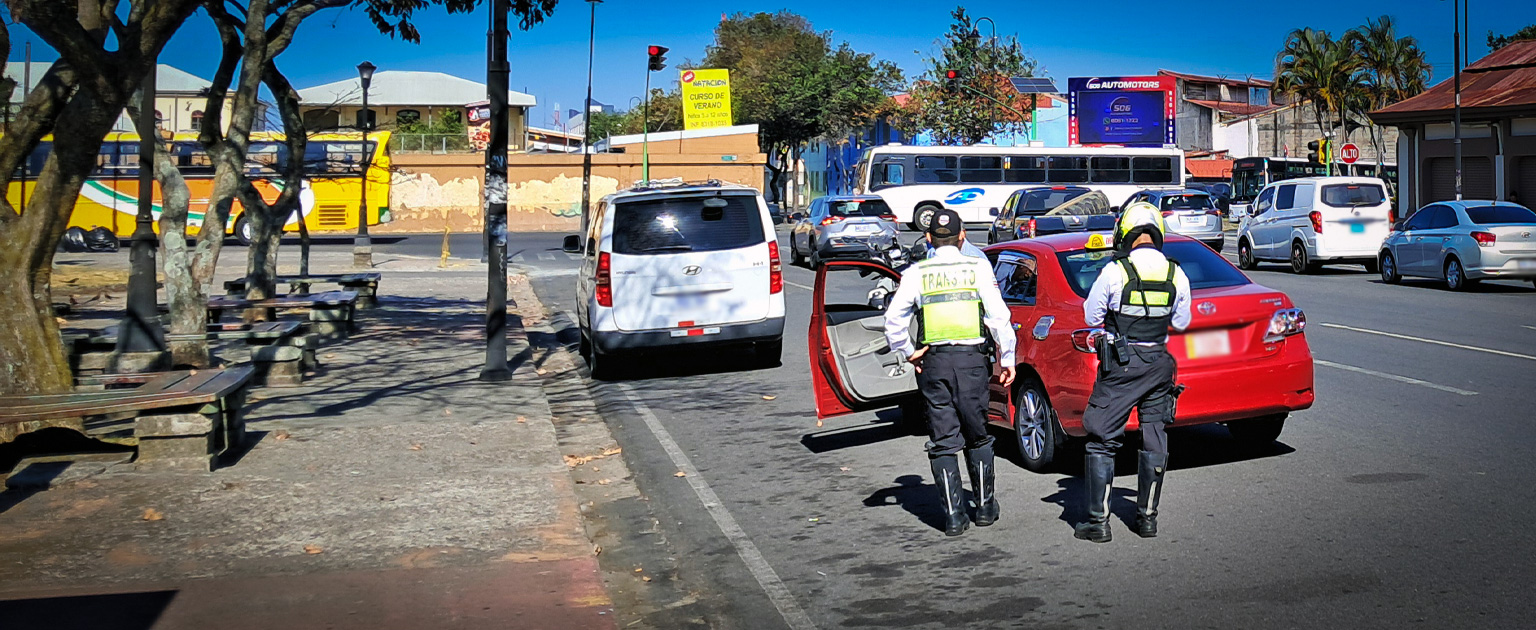 Oficiales de tránsito realizan control vehicular a conductor de taxi en una vía urbana con tráfico y buses al fondo