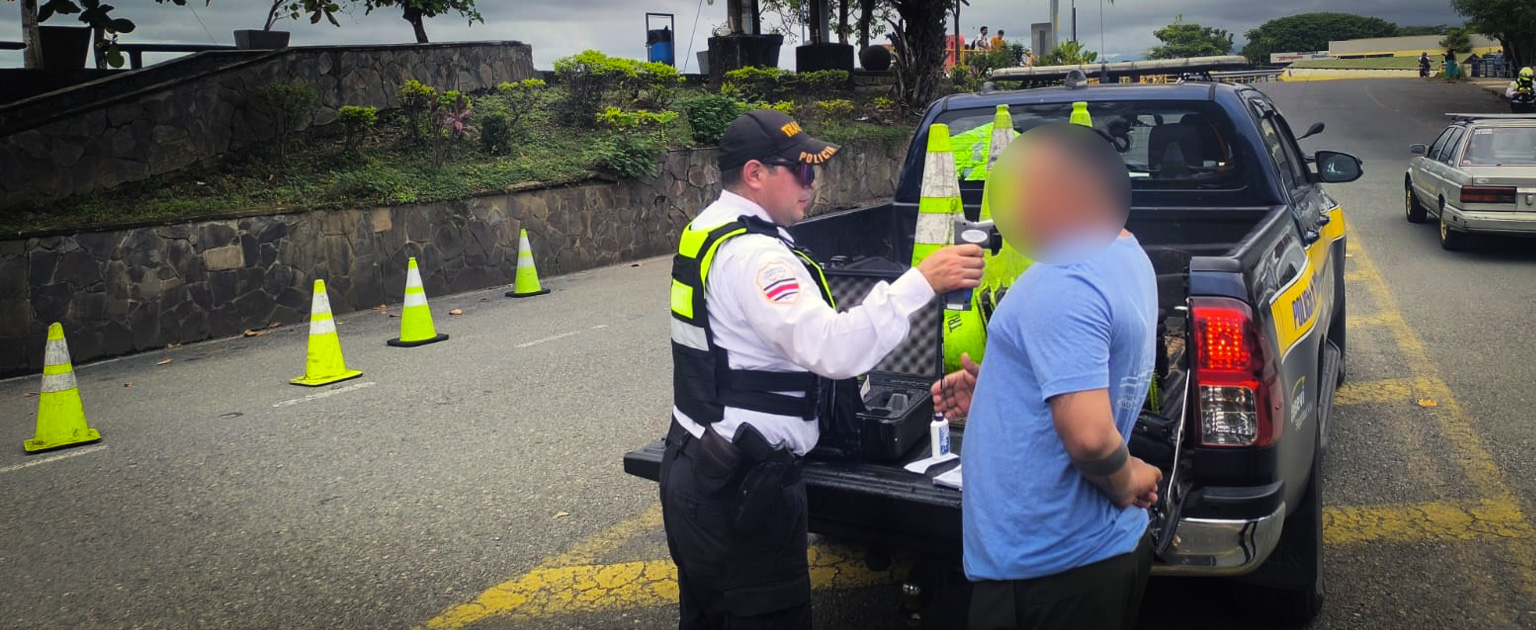 Persona oficial de tránsito realiza una prueba de alcoholemia a una persona conductora a un costado de una patrulla en la vía. En la carretera se observan conos de señalización que delimitan un operativo de control vehicular. Al fondo se aprecia una vía urbana y áreas ajardinadas junto a un muro de piedra.