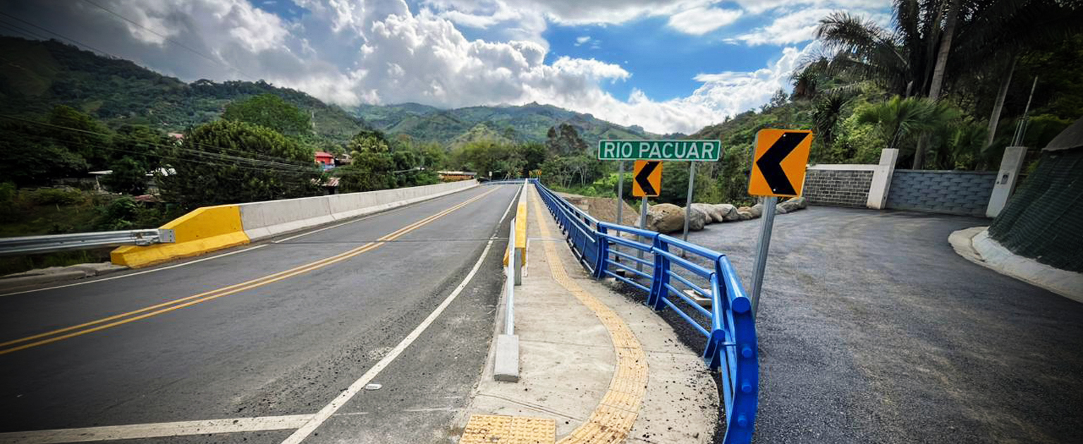 Puente vehicular sobre el río Pacuar en zona rural, con barandas azules y acera con señalización táctil. La vía asfaltada de dos carriles se extiende hacia un paisaje montañoso con abundante vegetación; a la derecha se observa un rótulo verde que indica “RÍO PACUAR” y señales de curva