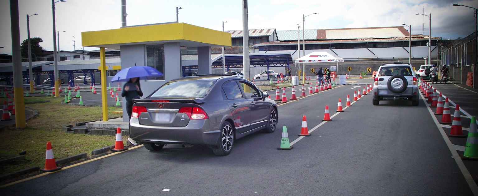 Circuito interno de pruebas prácticas, vehículos circulan en fila por un carril delimitado con conos; al lado se observa una caseta amarilla, señalización temporal y personal organizando las pruebas.