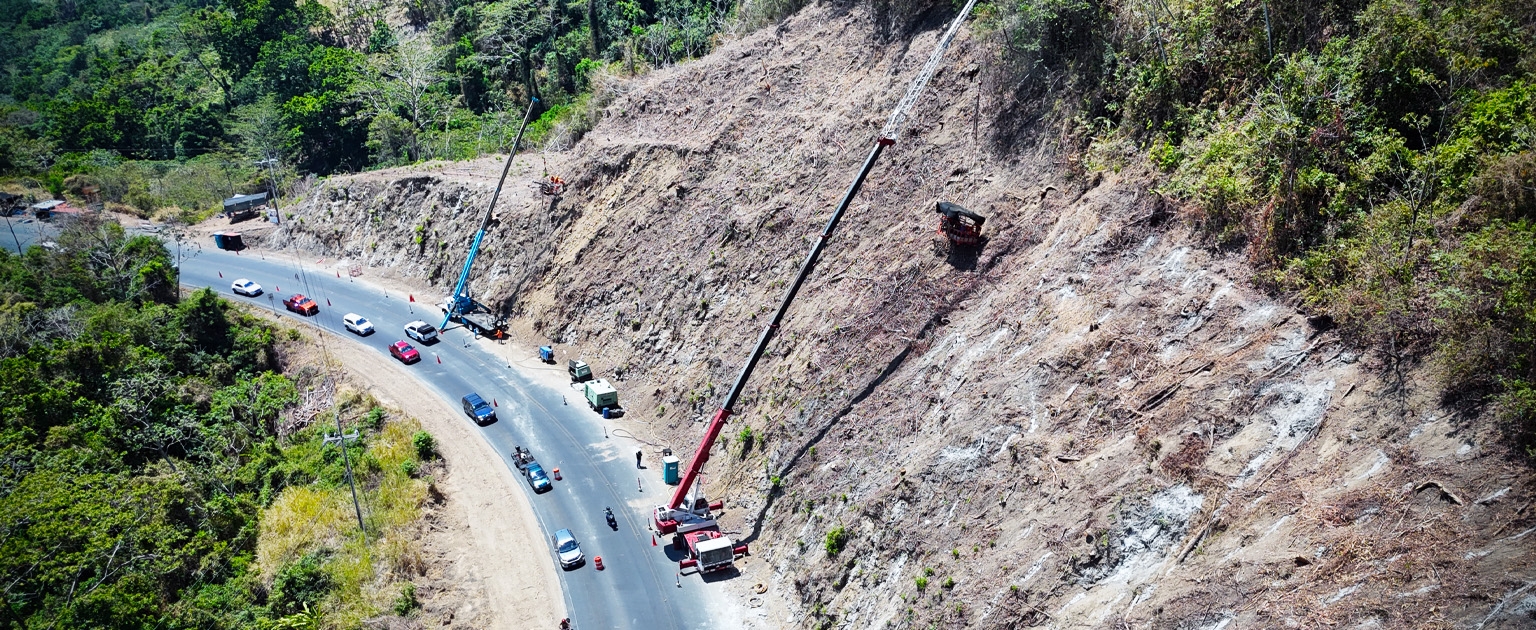 Vista aérea de trabajos de estabilización en un talud de la RN 34, sector de Caletas junto a una carretera, con grúas, maquinaria y tránsito vehicular controlado
