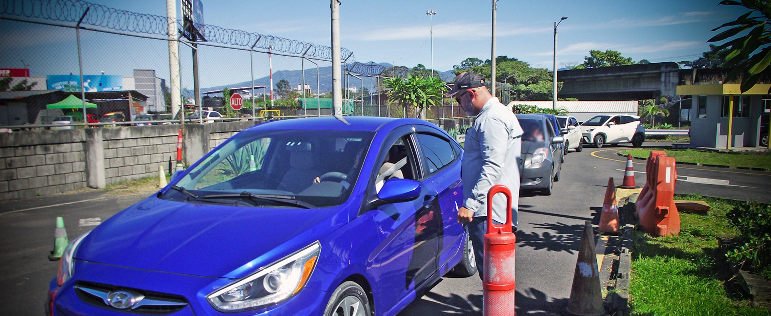 Persona realiza la prueba práctica en un vehículo azul , se observa un evaluador, conos de tránsito y varios autos en fila esperando su turno