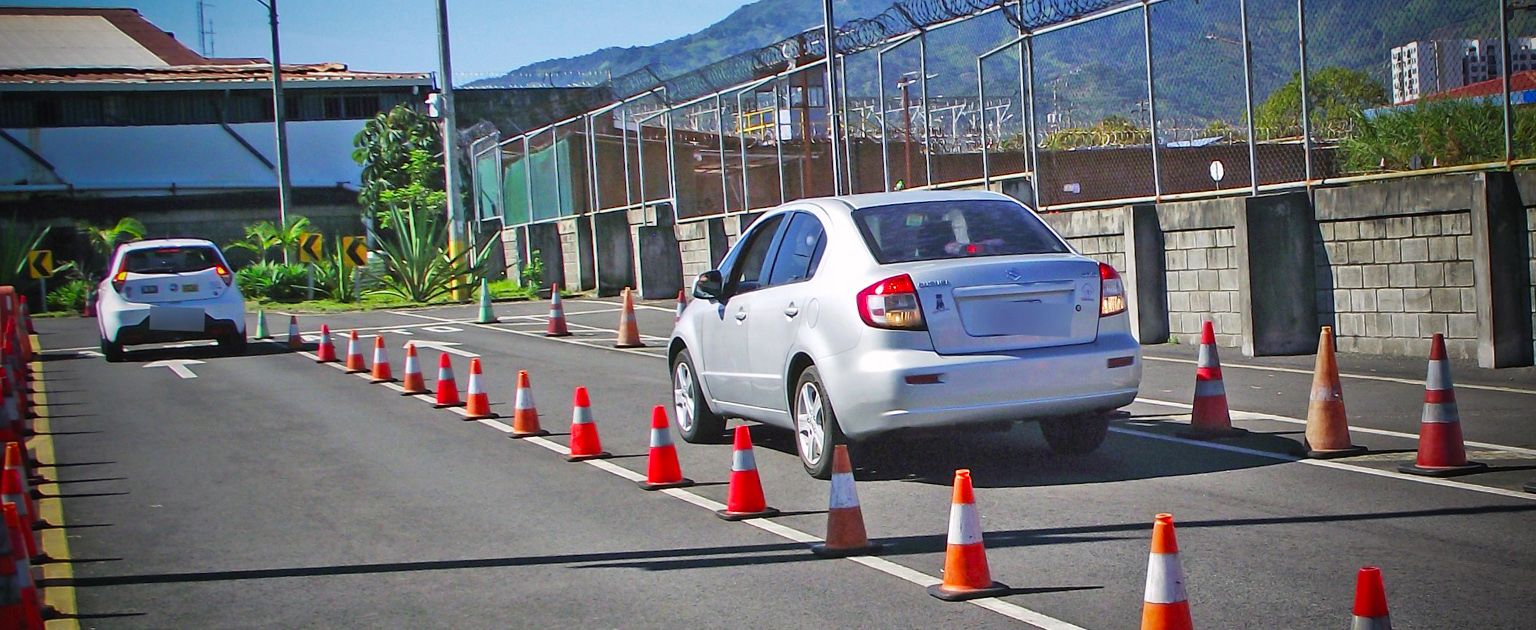 Fotografía muestra dos vehículos circulando por un tramo vial delimitado con conos de seguridad durante la pruebas práctica de manejo. La vía cuenta con carriles señalizados y está rodeada por cercas perimetrales, mientras al fondo se observan edificaciones y montañas bajo un cielo despejado.