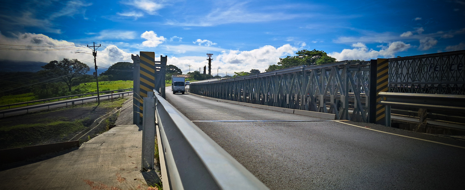 Fotografía del puente modular ubicado en las cercanías del Aeropuerto Internacional Juan Santamaría. La estructura metálica de un carril se extiende sobre una vía de acceso, mientras un camión circula por el puente. A ambos lados se observan barreras de seguridad, áreas verdes y postes eléctricos bajo un cielo azul con nubes blancas.
