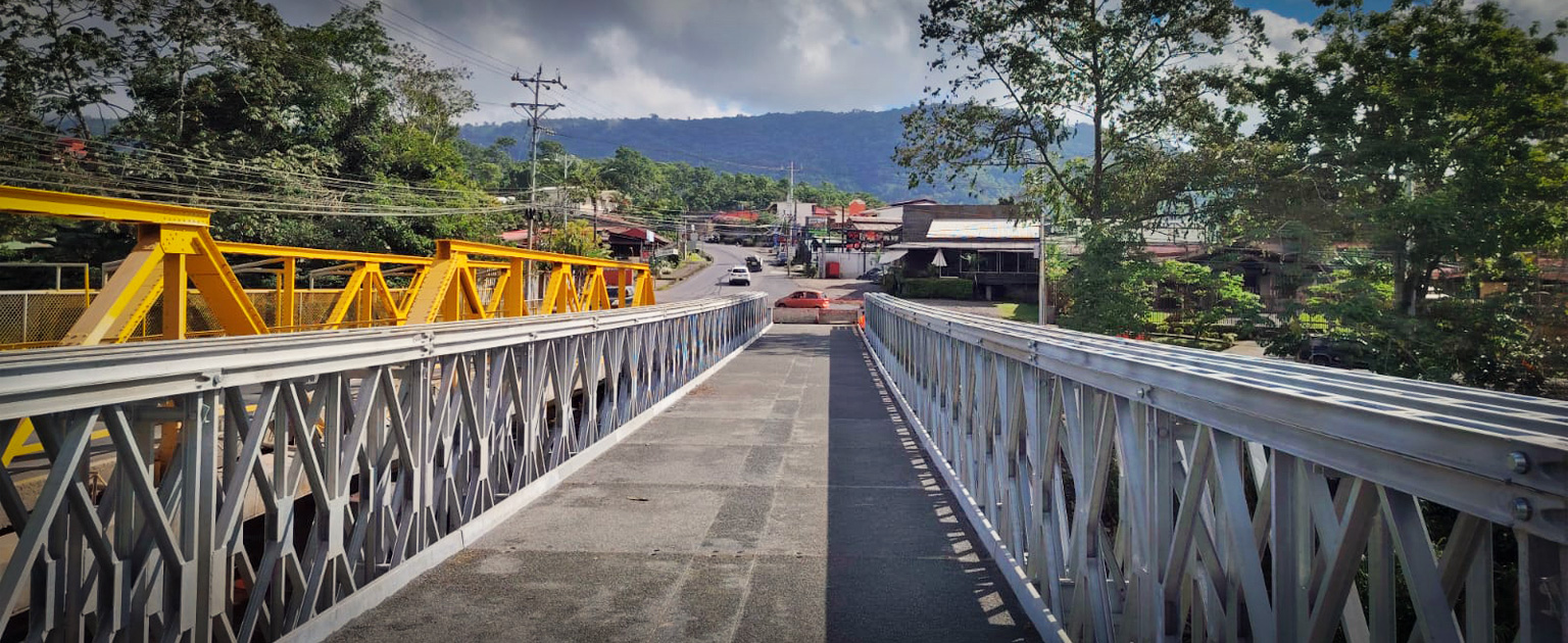 Vista sobre el nuevo puente modular instalado sobre el río Turrialba. La estructura metálica de color gris cuenta con barandales a ambos lados y se extiende hacia una zona urbana rodeada de vegetación. A la izquierda se observa el antiguo puente de acero pintado de amarillo, mientras al fondo destacan viviendas, árboles y montañas bajo un cielo parcialmente nublado.