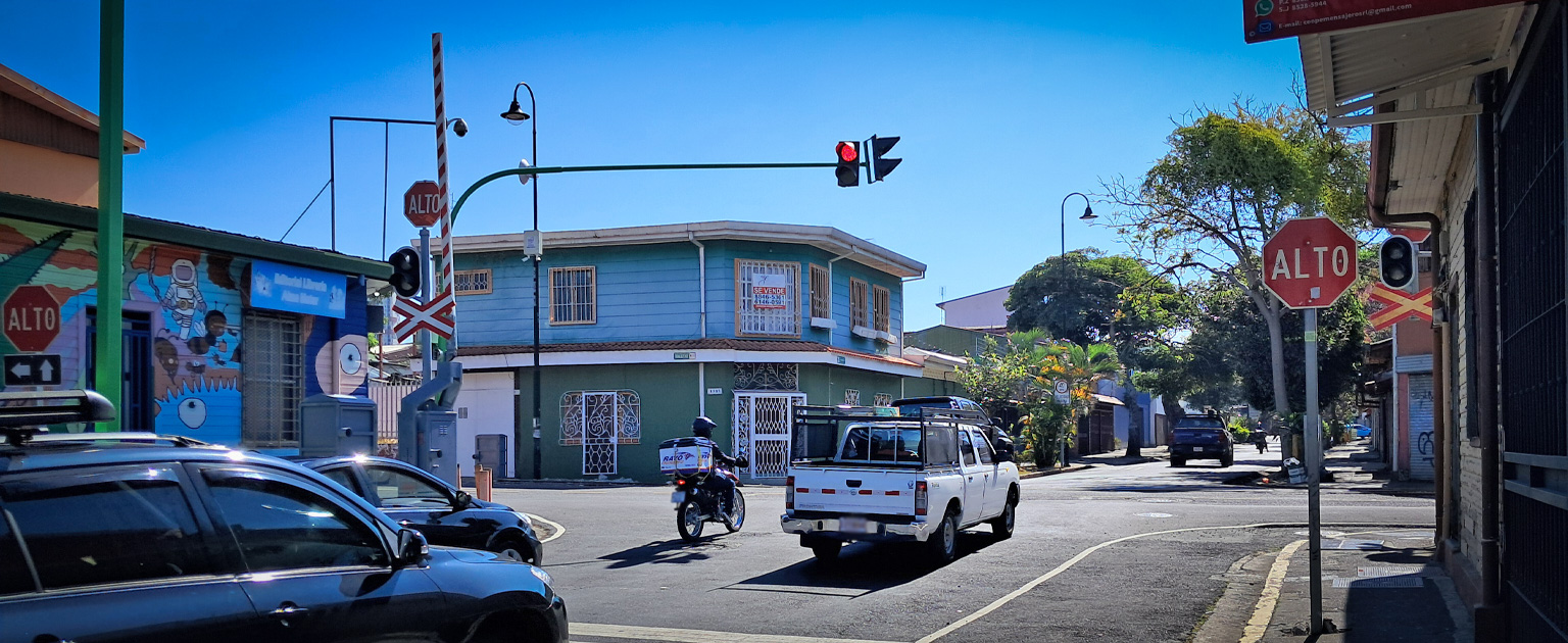 Intersección urbana con semáforo en rojo y señal de “ALTO”, donde circulan vehículos, incluyendo una motocicleta y un pick-up, rodeada de viviendas, señalización vial y vegetación en un entorno residencial.