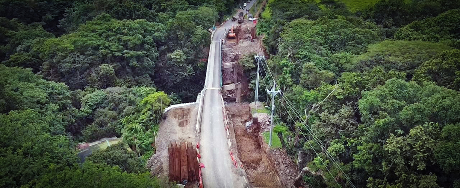 Vista aérea de los trabajos de construcción del puente sobre el río Tenorio. Se observa la carretera atravesando una zona boscosa, con maquinaria pesada y áreas de excavación en uno de los accesos del puente, donde se realizan obras de ampliación y mejora de la infraestructura vial.