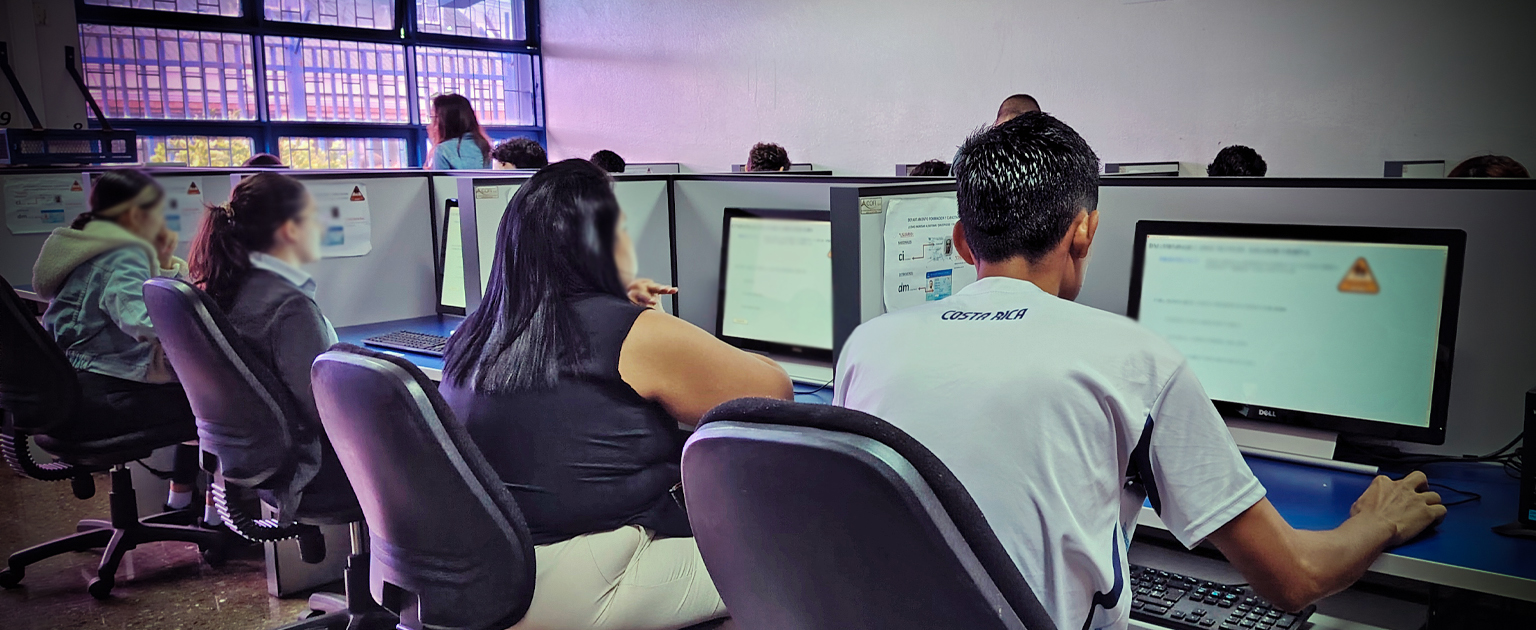 Personas sentadas frente a computadoras en un aula de evaluación, realizando pruebas teóricas; el espacio cuenta con filas de equipos, iluminación natural desde ventanas y supervisión al fondo.
