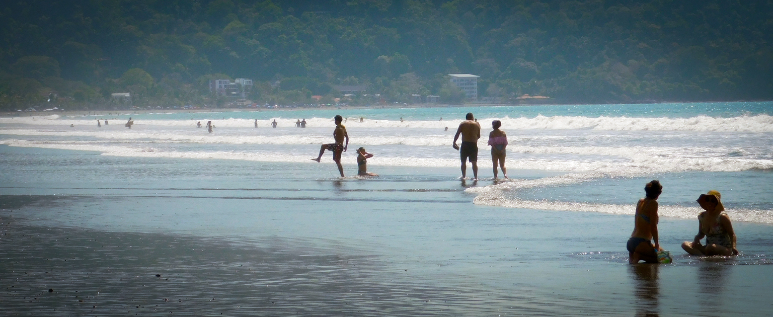 Personas disfrutan de una playa de amplia franja de arena mientras las olas llegan a la orilla. Al fondo se observan el mar y una zona montañosa cubierta de vegetación